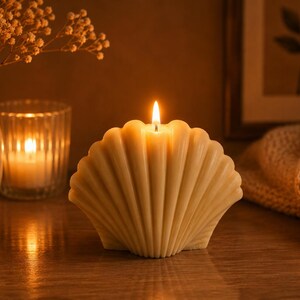 May include: A cream-colored, shell-shaped pillar candle with a lit wick. The candle features a distinct ribbed, fan-like texture resembling a scallop shell. It stands upright, emitting a warm, steady flame that illuminates its smooth, wax surface.