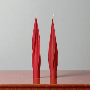May include: Two red spiral-shaped candles with a textured design. The candles have a tapered shape and a small, round base with a gold-colored band. The wicks are visible at the top. The candles are displayed on a wooden surface.