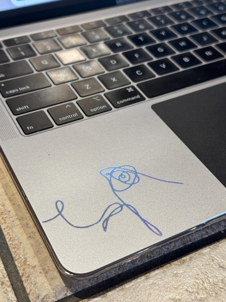 May include: A silver laptop computer with a blue and purple holographic sticker of a stylized rose on the top right corner of the trackpad.