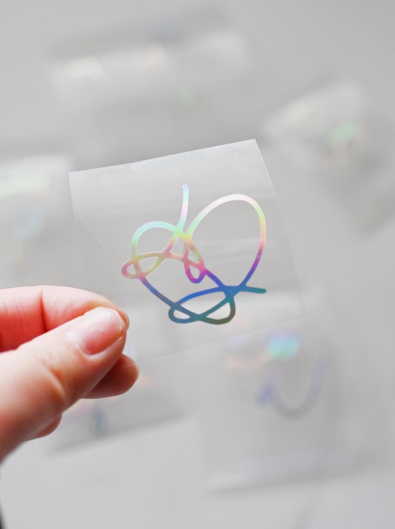 May include: A clear sheet of vinyl with a holographic iridescent heart-shaped design. The design is a stylized, abstract heart shape.