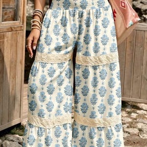 Può includere: Pantaloni a vita alta e gamba larga con base color crema e stampa floreale blu ripetuta. Il capo presenta una vita arricciata a punto smock e due fasce orizzontali di pizzo color crema sulle gambe, creando un effetto a balze stratificato.