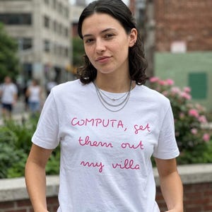 May include: A white short-sleeved t-shirt featuring the pink text "COMPUTA, get them out my villa" printed on the front. The shirt has a crew neckline and is worn with a layered silver chain necklace. The design is simple and casual in style.