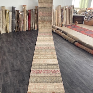 May include: A long, narrow, cream-colored rug with a repeating pattern of red, brown, and blue. The rug is rolled up and laid out on a wooden floor.