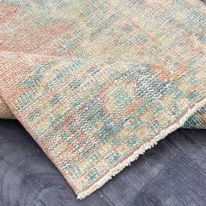 Könnte beinhalten: Ein gefalteter, abgenutzter Teppich mit verblasstem, Vintage-Aussehen. Der Teppich weist eine Mischung aus Farben auf, darunter Pfirsich, Türkis und Creme, mit einem strukturierten, gewebten Design. Die Kanten sind ausgefranst, was zur antiken Ästhetik des Teppichs beiträgt.