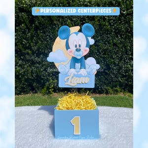 Peut inclure: Une bo&icirc;te de centre de table bleue avec un chiffre un jaune sur le devant. La bo&icirc;te est remplie de papier d&eacute;chiquet&eacute; jaune et surmont&eacute;e d'une d&eacute;coupe de Mickey Mouse, d'une lune et du nom "Liam". Le texte "PERSONALIZED CENTERPIECES" est en haut.