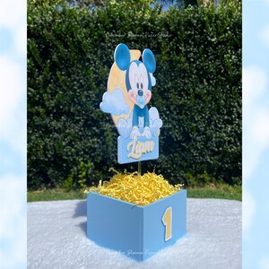 Peut inclure: Une bo&icirc;te bleue avec le chiffre 1 jaune, remplie de papier d&eacute;chiquet&eacute; jaune. Une figurine de Mickey Mouse avec le nom "Liam" est au-dessus, parfaite pour un premier anniversaire.