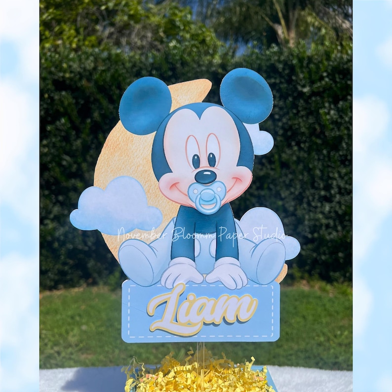Peut inclure: D&eacute;coupe de Mickey Mouse b&eacute;b&eacute; avec une sucette bleue, devant une lune jaune et des nuages blancs. Le nom "Liam" est &eacute;crit sur une pancarte bleue. Les mots "November Bloom Paper Studio" sont &eacute;galement visibles.