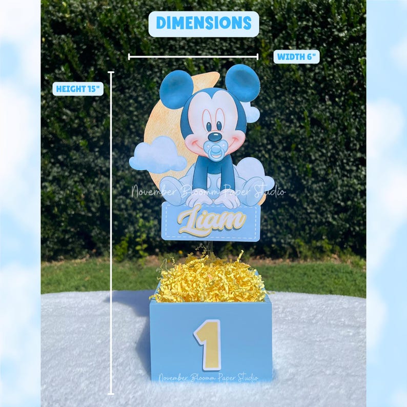 Peut inclure: Centre de table bleu clair avec une d&eacute;coupe de Mickey Mouse, une pancarte avec le nom "Liam" et le chiffre 1. Le centre de table mesure 38 cm de haut et 15 cm de large. La bo&icirc;te est remplie de papier d&eacute;chiquet&eacute; jaune.