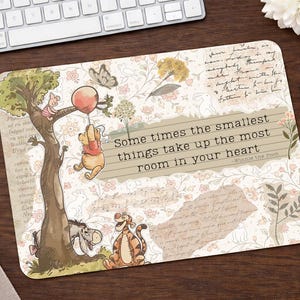 May include: A rectangular mouse mat with a Winnie the Pooh design. The design includes illustrations of Pooh, Piglet, Eeyore, and Tigger. The mouse mat has the quote, "Some times the smallest things take up the most room in your heart."
