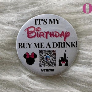 Puede incluir: Botón blanco con el texto "It's my Birthday Buy me a drink!" con un Mickey Mouse rosa, un código QR y un castillo rosa con una cabeza de Mickey Mouse en la parte superior. El botón también tiene la palabra "venmo" impresa.