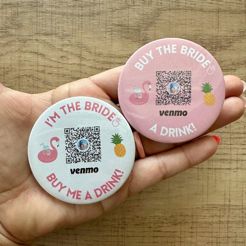Peut inclure: Deux boutons ronds roses et blancs avec un flamant rose, un ananas et un code QR. Le bouton rose dit "Achetez un verre &agrave; la mari&eacute;e !" et le bouton blanc dit "Je suis la mari&eacute;e, achetez-moi un verre !" Les deux boutons portent le texte "venmo".