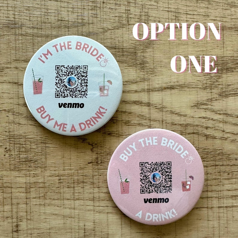Peut inclure: Deux boutons ronds, blanc et rose, avec un code QR, le texte "I'm the Bride" et "Buy me a drink!" et le mot "venmo" sur chacun. Les boutons sont sur une surface en bois.