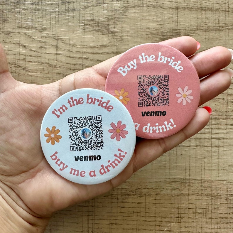 Peut inclure: Deux autocollants en papier ronds, roses et blancs, avec un code QR et le texte "I'm the bride buy me a drink!" et "Buy the bride a drink!" avec le mot "venmo" imprim&eacute; sous le code QR.