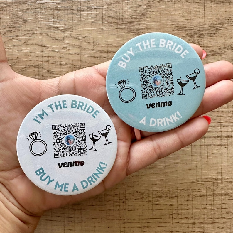 Peut inclure: Deux dessous-de-verre ronds, blancs et bleus avec un code QR, une bague et deux verres &agrave; martini. Le texte sur les dessous-de-verre est "I'm the Bride, Buy me a drink!" et "Buy the Bride, A Drink!" avec le mot "venmo" imprim&eacute; sous le code QR.