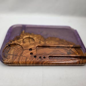 May include: A rectangular wooden tray with a purple resin border. The tray features a natural wood grain pattern with circular and rectangular cutouts. The tray is designed for organizing small items.