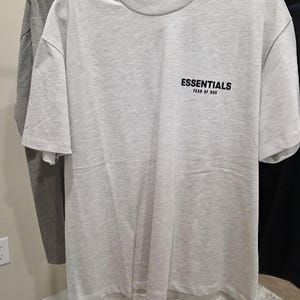 May include: A light gray t-shirt with the word "ESSENTIALS" and "FEAR OF GOD" printed in black on the front. The shirt is hanging on a white hanger. The shirt has short sleeves and a crew neck.