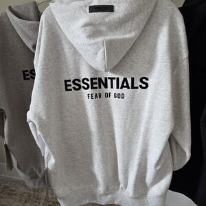 Op de afbeelding: Een lichtgrijze hoodie met het woord "ESSENTIALS" in het zwart op de rug gedrukt, met "FEAR OF GOD" eronder. De hoodie heeft een capuchon en een klein zwart label aan de bovenkant van de capuchon. Andere hoodies zijn zichtbaar op de achtergrond.
