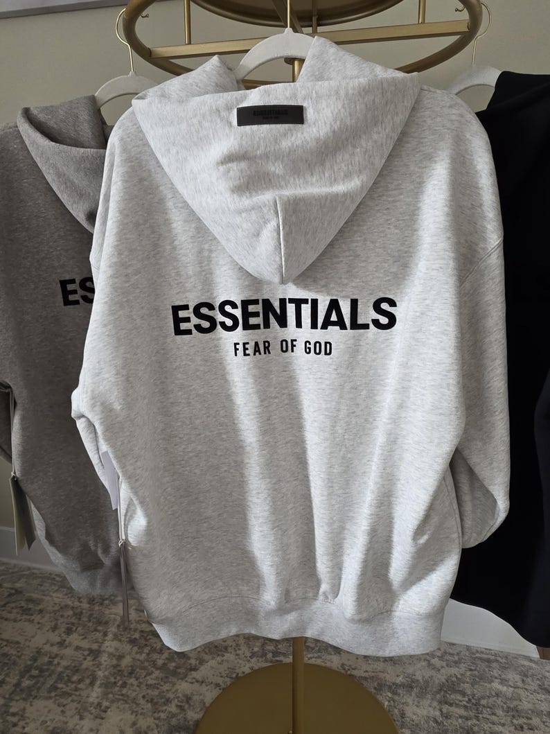 Op de afbeelding: Een lichtgrijze hoodie met het woord "ESSENTIALS" in het zwart op de rug gedrukt, met "FEAR OF GOD" eronder. De hoodie heeft een capuchon en een klein zwart label aan de bovenkant van de capuchon. Andere hoodies zijn zichtbaar op de achtergrond.