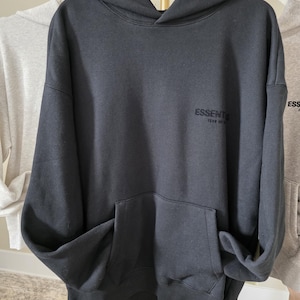 Op de afbeelding: Een donkergrijze hoodie met een kangoeroezak aan de voorkant en een capuchon. Het woord "ESSENTIALS" staat op de borst gedrukt. De hoodie hangt aan een witte hanger.