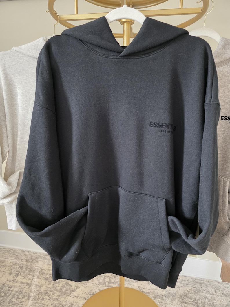 Op de afbeelding: Een donkergrijze hoodie met een kangoeroezak aan de voorkant en een capuchon. Het woord "ESSENTIALS" staat op de borst gedrukt. De hoodie hangt aan een witte hanger.