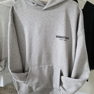 Op de afbeelding: Een grijze hoodie met een voorzak en een capuchon. Het woord "ESSENTIALS" is in het zwart op de voorkant gedrukt, met "FEAR OF GOD" eronder. De hoodie hangt aan een witte hanger.