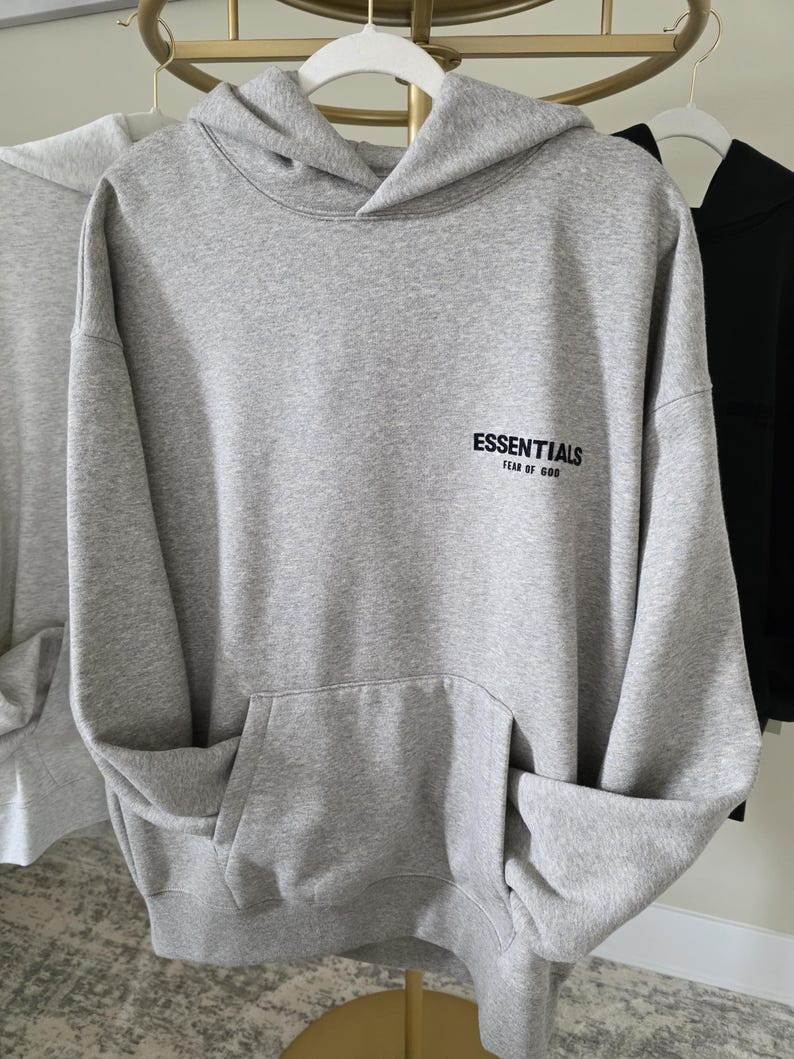 Op de afbeelding: Een grijze hoodie met een voorzak en een capuchon. Het woord "ESSENTIALS" is in het zwart op de voorkant gedrukt, met "FEAR OF GOD" eronder. De hoodie hangt aan een witte hanger.