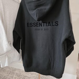 Op de afbeelding: Zwarte hoodie met het woord "ESSENTIALS" en "FEAR OF GOD" op de rug gedrukt. De hoodie heeft een capuchon en een label. Een andere grijze hoodie hangt aan de linkerkant.