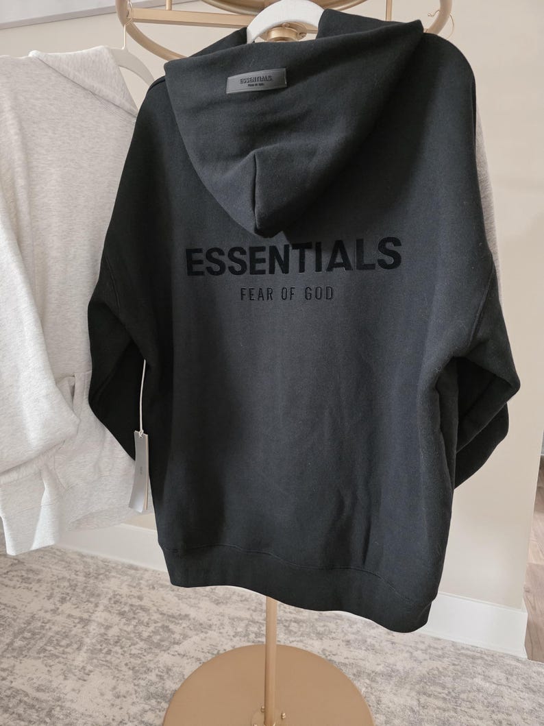 Op de afbeelding: Zwarte hoodie met het woord "ESSENTIALS" en "FEAR OF GOD" op de rug gedrukt. De hoodie heeft een capuchon en een label. Een andere grijze hoodie hangt aan de linkerkant.
