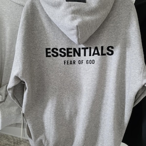 Op de afbeelding: Een grijze hoodie met het woord "ESSENTIALS" in zwarte letters op de rug, met "FEAR OF GOD" eronder. De hoodie heeft een capuchon en een klein zwart label op de capuchon.