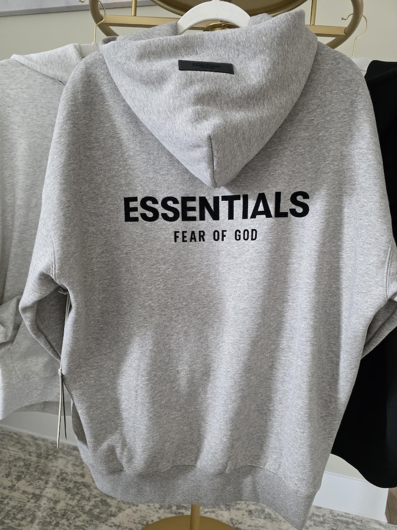 Op de afbeelding: Een grijze hoodie met het woord "ESSENTIALS" in zwarte letters op de rug, met "FEAR OF GOD" eronder. De hoodie heeft een capuchon en een klein zwart label op de capuchon.