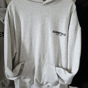 Op de afbeelding: Een lichtgrijze hoodie met een kangoeroezak en een capuchon. Het woord "ESSENTIALS" is in het zwart op de voorkant gedrukt. De hoodie hangt aan een witte hanger.