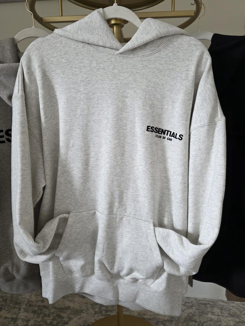 Op de afbeelding: Een lichtgrijze hoodie met een kangoeroezak en een capuchon. Het woord "ESSENTIALS" is in het zwart op de voorkant gedrukt. De hoodie hangt aan een witte hanger.