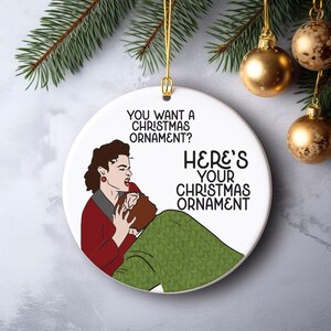 May include: A round white ceramic Christmas ornament featuring a graphic illustration of a person in a red jacket grabbing the head of another person in a green sweater. The text reads: YOU WANT A CHRISTMAS ORNAMENT? HERE'S YOUR CHRISTMAS ORNAMENT.