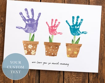May include: A white card with three handprint flowers in pots. The flowers are purple, pink, and blue. The text "we love you so much mommy" is written below the flowers.