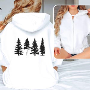 May include: White zippered hoodie with a black silhouette of a row of pine trees on the back. The hoodie is shown on a model.