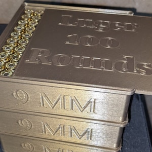 May include: A stack of four gold-coloured boxes with a lid. The lid is open and has the text "Luger 100 Rounds" on it. The boxes are labelled "9MM".