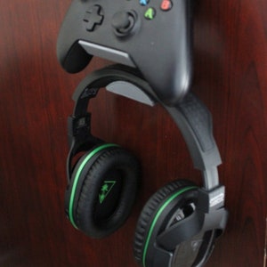 May include: A black gaming headset with green accents hangs on a wall mount next to a black video game controller.
