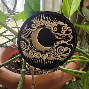 May include: A round, black embroidered patch featuring a crescent moon and sunburst design in gold thread. The patch is surrounded by stylized clouds and sun rays. The patch is set against a backdrop of green leaves and a terracotta pot.