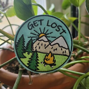 May include: Round embroidered patch with the words "GET LOST" above a mountain scene. The patch features green pine trees, a yellow campfire, and a rising sun. The patch is on a light blue background.