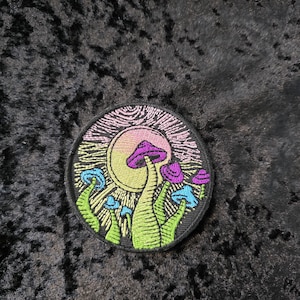 May include: Round embroidered patch featuring a colorful mushroom design. The patch has a black border and depicts mushrooms in shades of purple, blue, and green, with a yellow sun in the background. The design is detailed and vibrant.