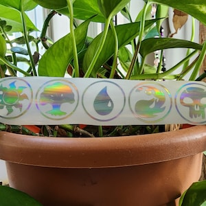 May include: A white sticker with five iridescent holographic circles. Each circle contains a different symbol: a sun, a tree, a teardrop, a wave, and a skull.