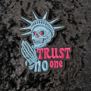 May include: Embroidered patch featuring a skull with the Statue of Liberty's crown, praying hands, and the text "TRUST NO ONE" in pink and blue. The patch is on a black background.
