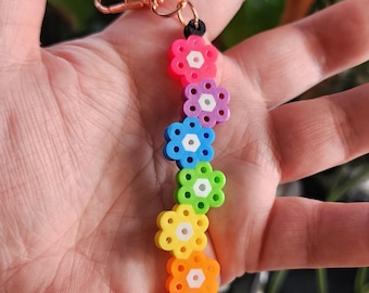 Puede incluir: Un llavero de cuentas de colores arcoíris con un diseño de flores. Cada flor es de un color diferente del arcoíris, con un centro blanco. El llavero tiene un cierre dorado.