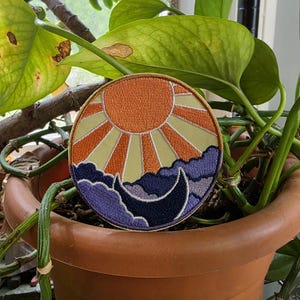 May include: A circular embroidered patch featuring a vibrant orange sun with radiating beams against a pale yellow sky. Below the sun, a dark purple crescent moon sits nestled among stylized, layered purple clouds with a gold border.