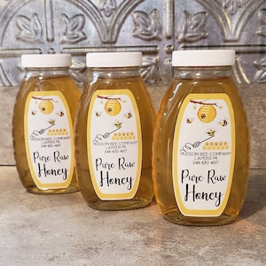 Op de afbeelding: Drie glazen potten gevuld met honing, elk met een geel etiket waarop "Pure Raw Honey" staat en de bedrijfsnaam "Hudson Bee Company" en contactgegevens. De etiketten bevatten ook een cartoonillustratie van een bijenkorf met bijen.