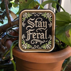 May include: A rectangular embroidered patch with a black background and a gold border. The text "Stay Feral" is stitched in white in the center, surrounded by a decorative frame featuring light green leaves and small white flowers at the corners.
