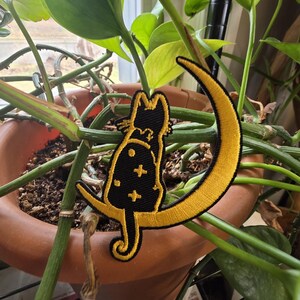 May include: A black cat patch with gold trim sits on a gold crescent moon. The cat has a small bow and is decorated with gold stars. The patch is set against a backdrop of green leaves and a brown pot.