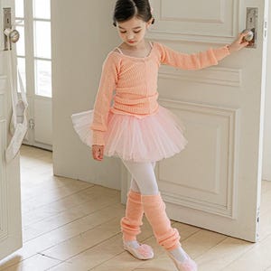 May include: A young girl in a ballet outfit, featuring a peach-colored long-sleeved sweater, a pink tutu, white leggings, and matching leg warmers. She wears ballet slippers with a flower accent. The outfit is designed for dance and performance.