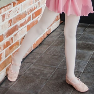 May include: A young person wearing pink ballet shoes and white tights, standing on a tiled floor with a brick wall in the background.