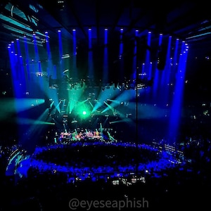 May include: A concert stage with blue and green lights illuminating a large crowd of people. The stage is set up with a large screen and a band is performing. The text "@eyeseaphish" is visible at the bottom of the image.